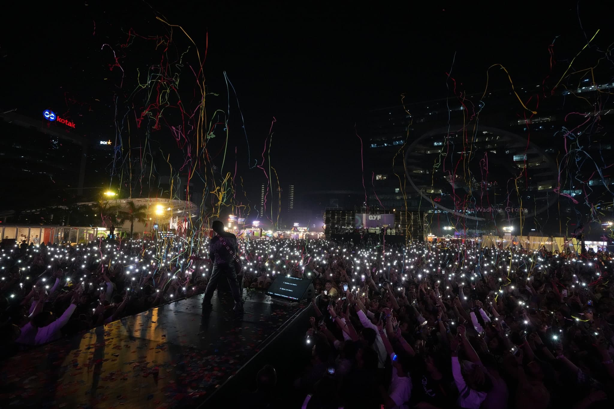 A concert performer faces a crowd lit by phone lights and falling streamers